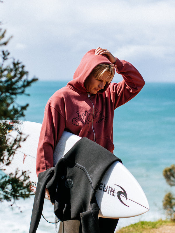Person wearing a maroon hoodie with a logo, holding a surfboard with a visible brand logo, standing on a grassy area with ocean in the background.