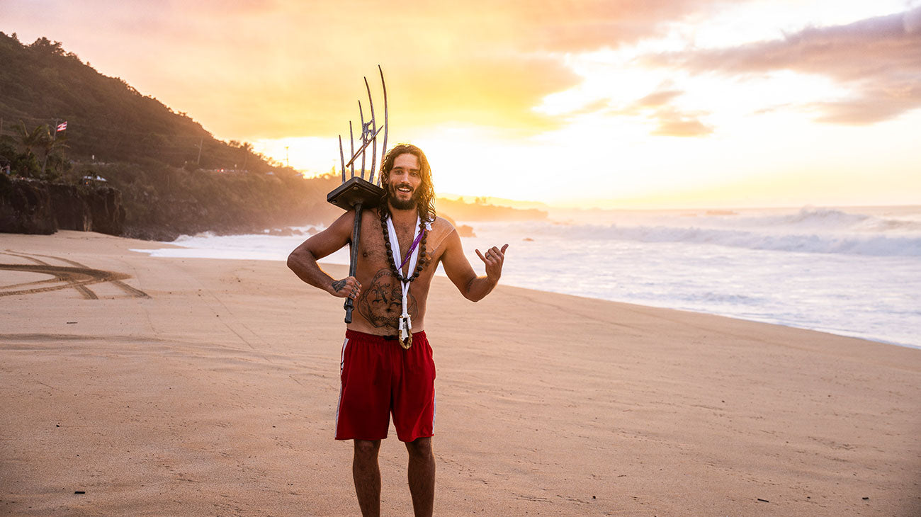 Man with tattoos and red shorts standing on a beach at sunset