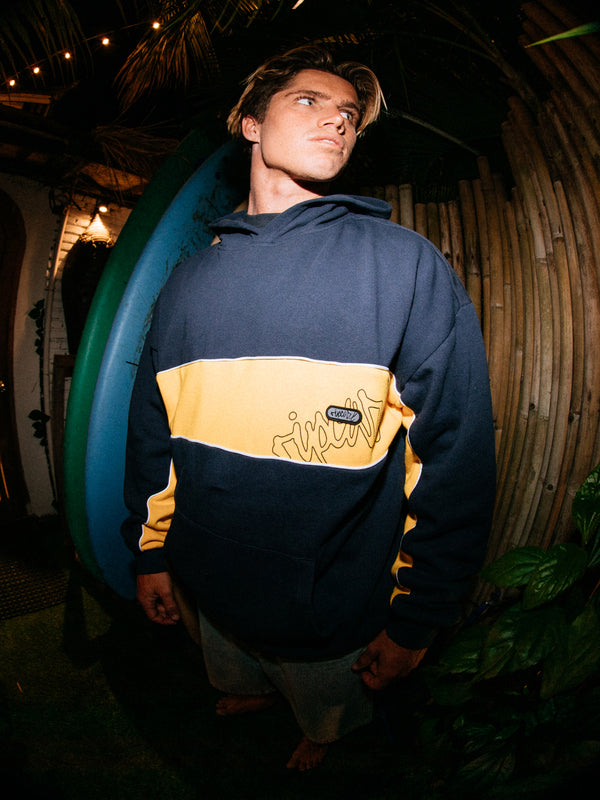 Person wearing a blue and yellow hoodie with a logo, standing in a tropical setting.