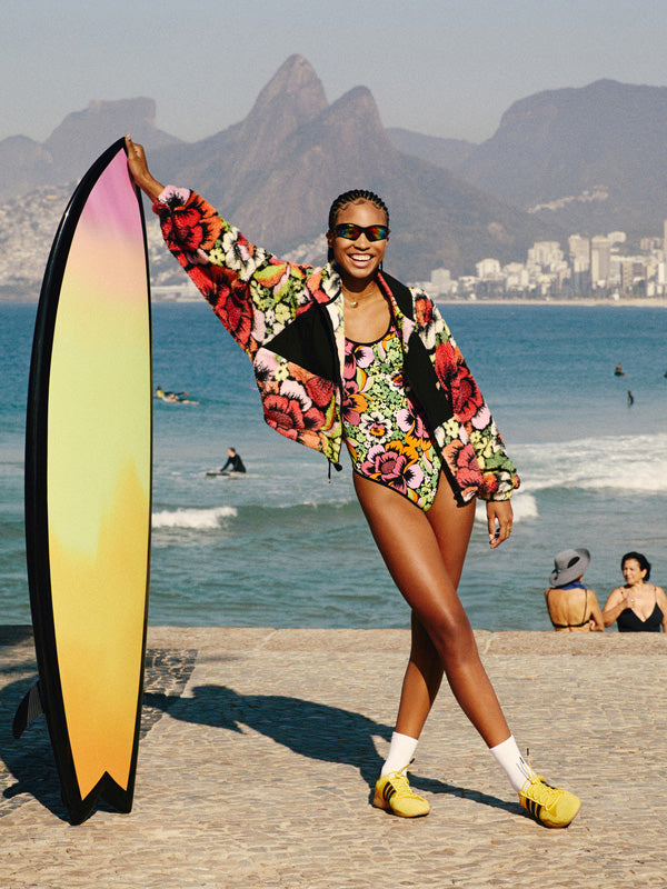 Woman in a colorful outfit holding a surfboard on a beach with mountains in the background