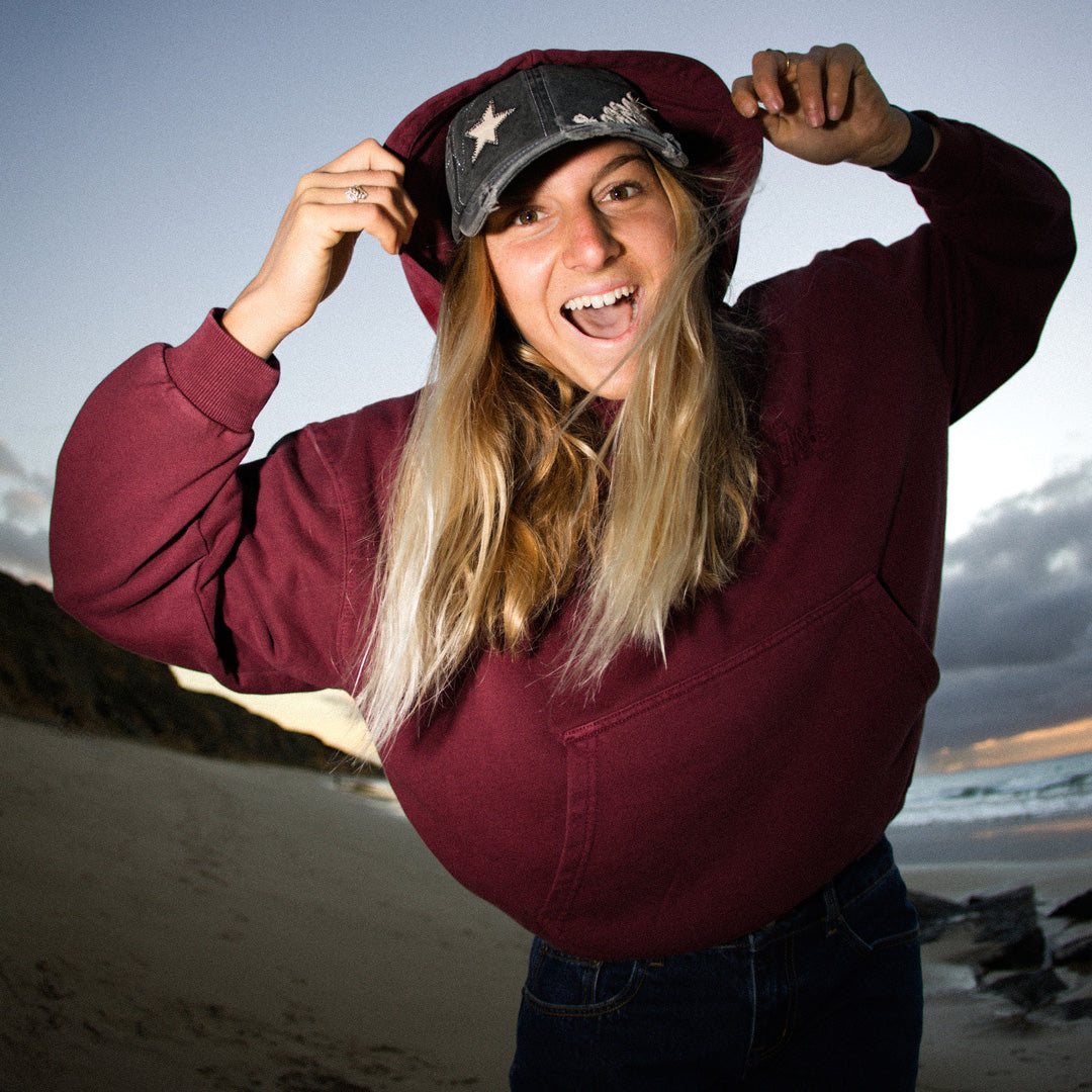 Person wearing a maroon hoodie and cap on a beach with a cloudy sky.