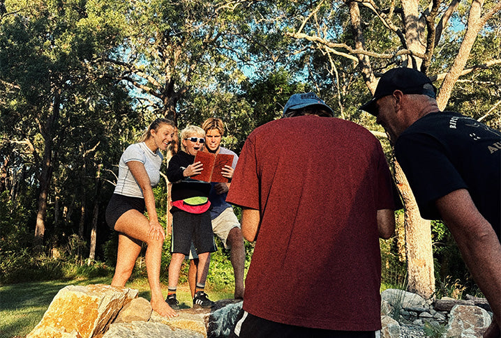 Group of people outdoors, with one person reading a book, surrounded by trees and rocks.
