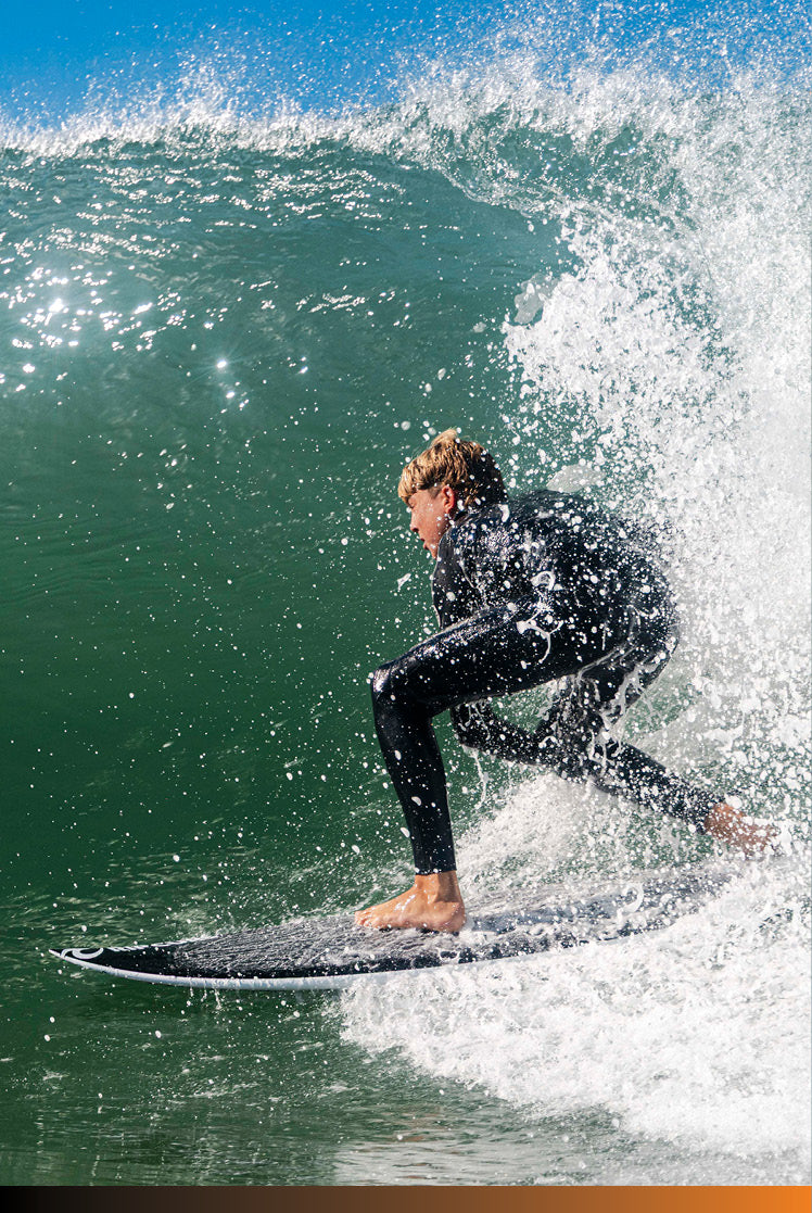 A surfer in a black wetsuit crouches on a surfboard inside a curling ocean wave, with white spray and blue sky visible.