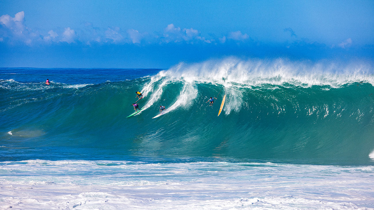 Surfers riding a large wave in the ocean with a clear blue sky.