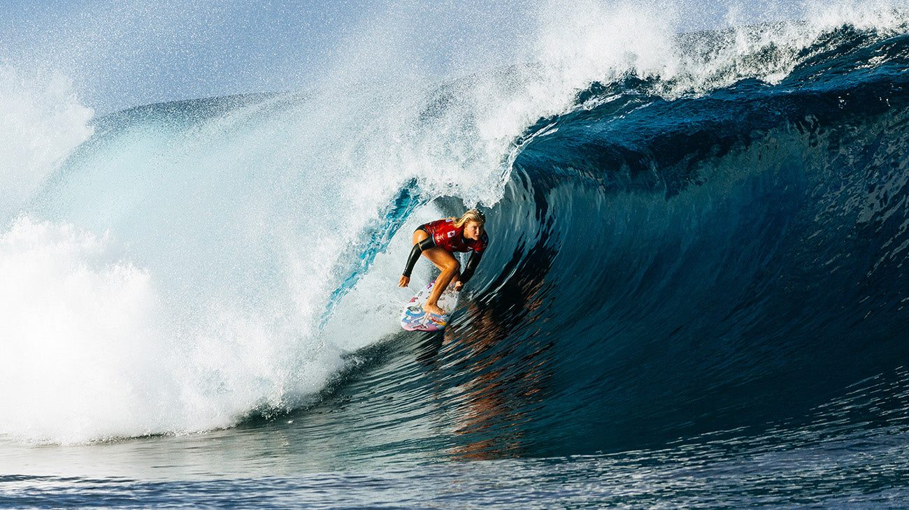 A surfer in a red and black wetsuit rides inside the barrel of a large blue ocean wave with white spray, captured in action.