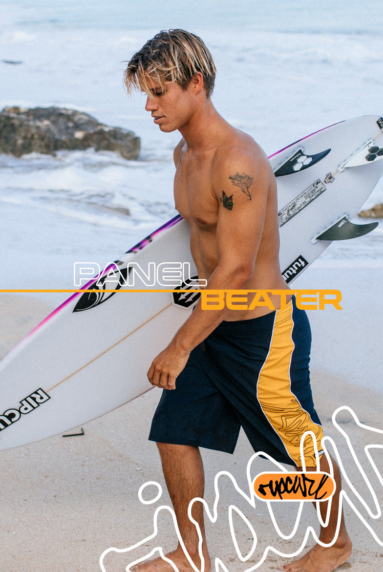 Man holding a surfboard on a beach with 'Panel Beater' branding.