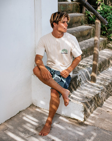 Man sitting on stone steps wearing a white t-shirt with a logo and green shorts.