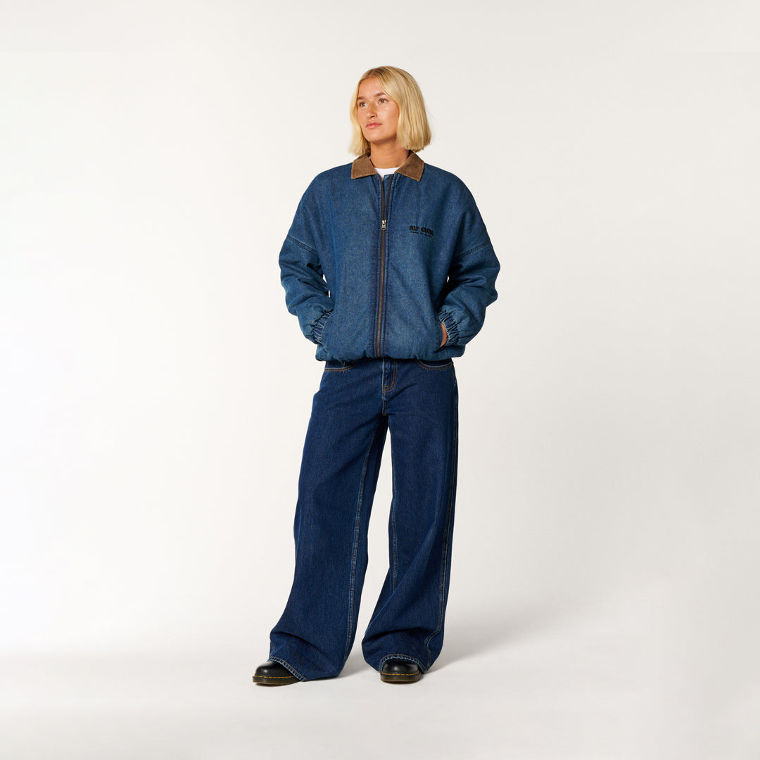 A model stands against a plain light gray studio background wearing a blue denim jacket with a brown collar and matching wide-leg blue denim pants.