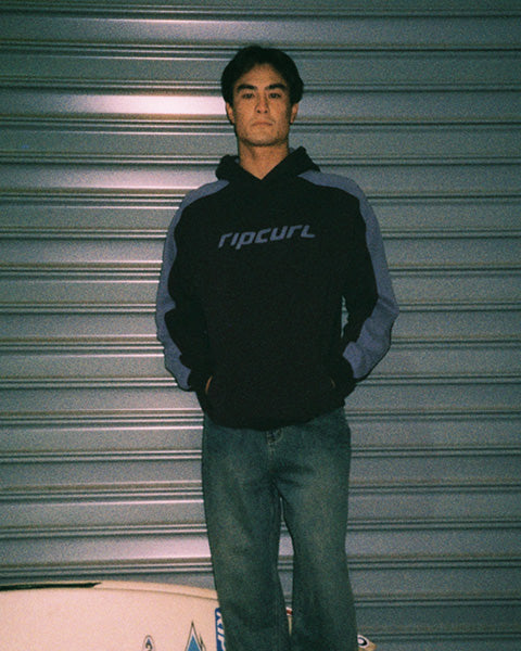 A man wearing a black and gray Rip Curl hoodie with blue jeans, standing in front of a gray metal roll-up door.