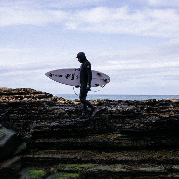 A surfer in a black wetsuit carrying a white surfboard walks along dark rocky shoreline under a cloudy sky.