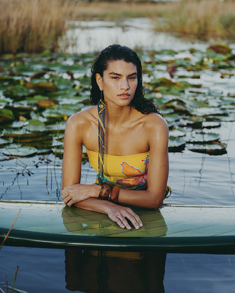 A woman posing on a green surfboard in a lily-patched lake, wearing a yellow strapless top with a colorful butterfly print and brown leather bracelets.