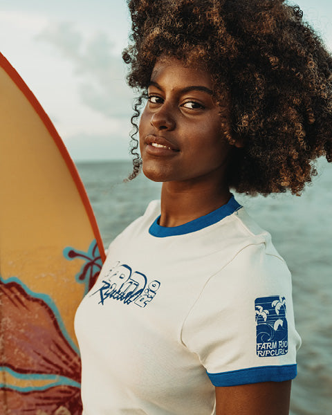 A woman with an afro hairstyle wearing a white and blue surf-themed t-shirt, standing next to a yellow surfboard with the ocean in the background.