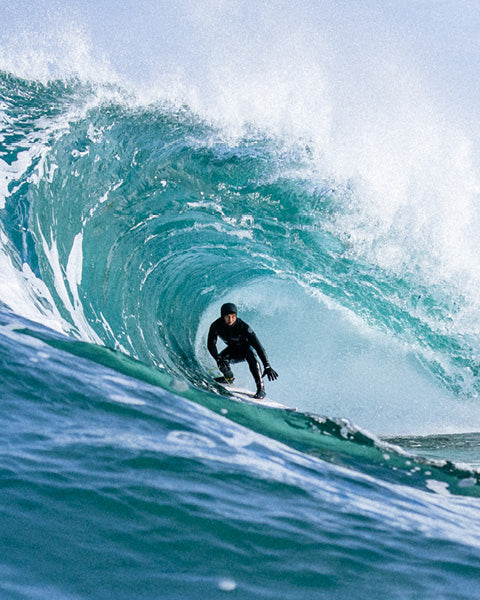 A surfer in a black wetsuit rides inside the curl of a large turquoise ocean wave with white spray, photographed from a low angle.