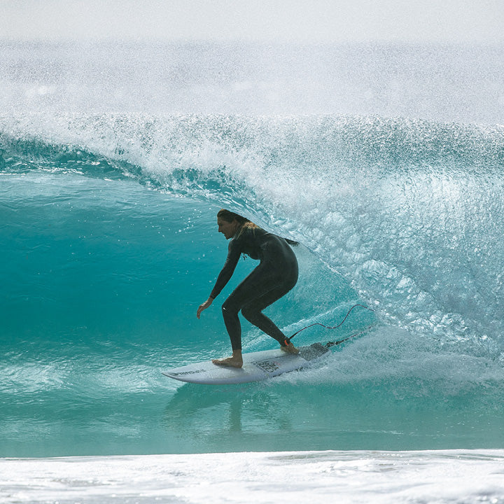 A surfer in a black wetsuit rides a turquoise ocean wave on a white surfboard, with white spray and foam around them.
