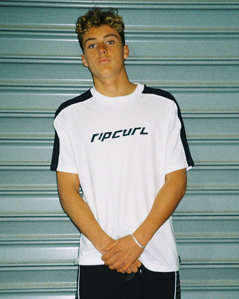 A young man wearing a white Rip Curl t-shirt with black shoulder panels and black shorts, standing in front of a gray metal shutter.