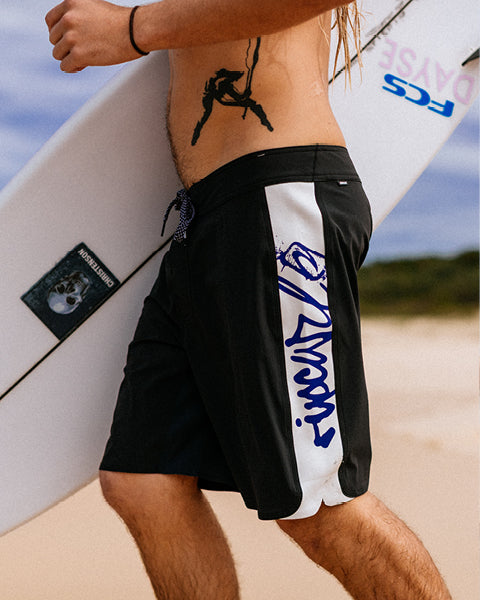 Person wearing black and white swim shorts with a logo, standing on a beach with a surfboard.
