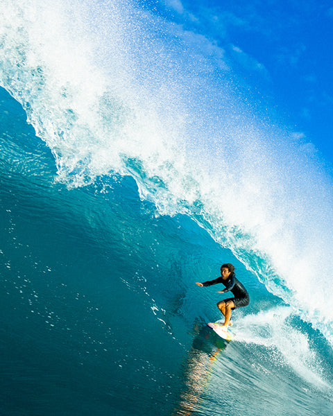 Surfer riding a large wave in clear blue water with a bright blue sky.