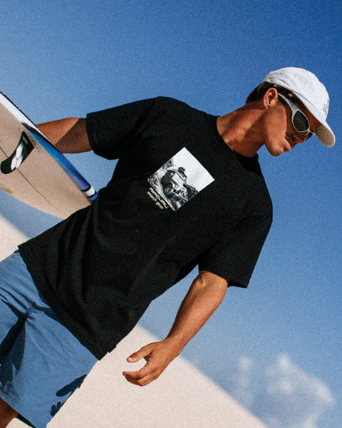 Person wearing a black t-shirt with a graphic design, blue shorts, white cap, and sunglasses against a clear blue sky.