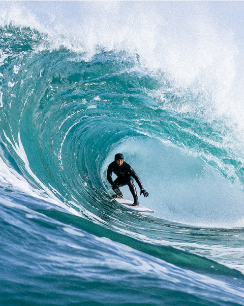 A surfer in a black wetsuit rides inside the barrel of a large turquoise ocean wave with white spray.