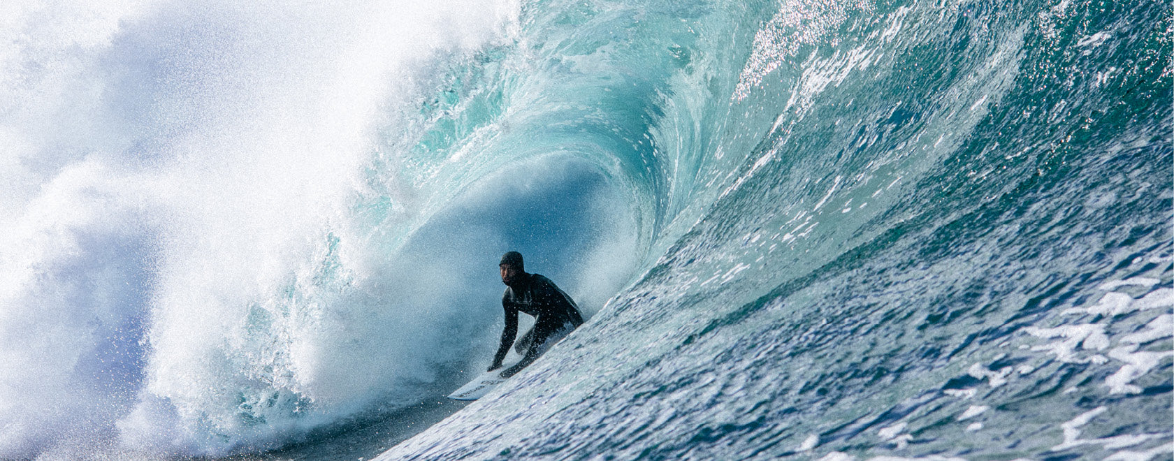 A surfer in a black wetsuit rides inside the curl of a large turquoise ocean wave with white spray, photographed from a low angle.