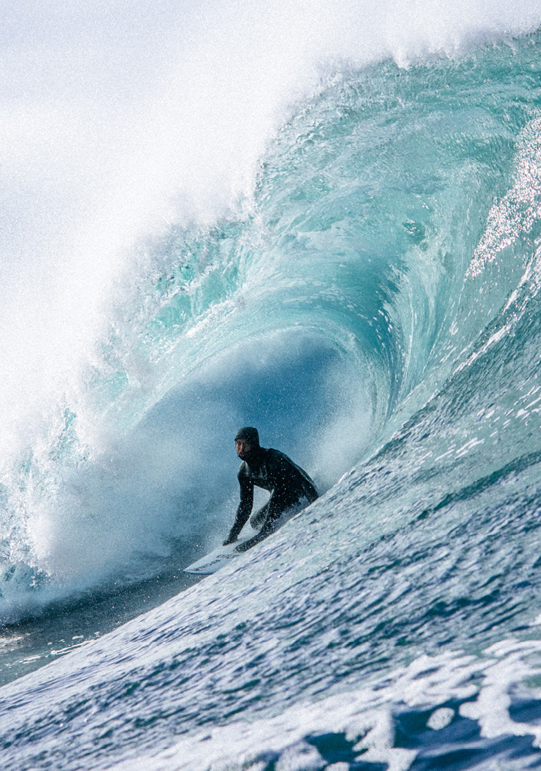 A surfer in a black wetsuit rides inside the barrel of a large turquoise ocean wave with white foam curling around it.