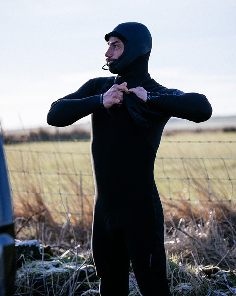 Person wearing a black wetsuit standing in a field with a fence and sky in the background
