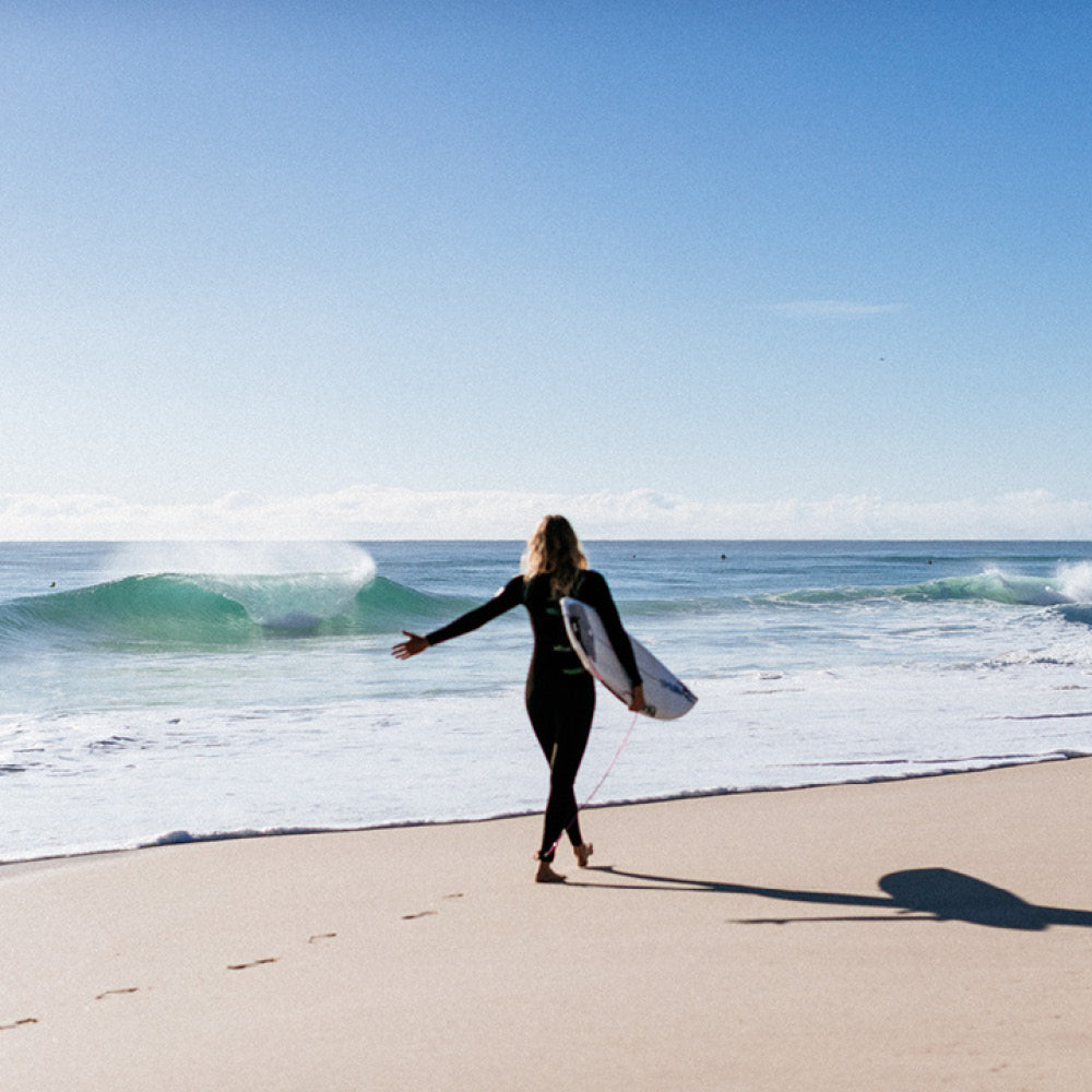 Person in a wetsuit holding a surfboard on a beach with waves in the background
