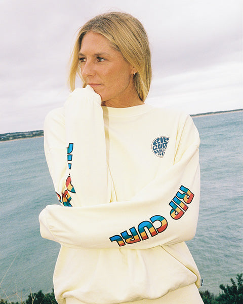 Person wearing a white sweatshirt with colorful text and graphics, standing by a body of water.