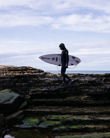 A surfer in a black wetsuit carrying a white surfboard walks along a rocky shoreline under a cloudy sky.
