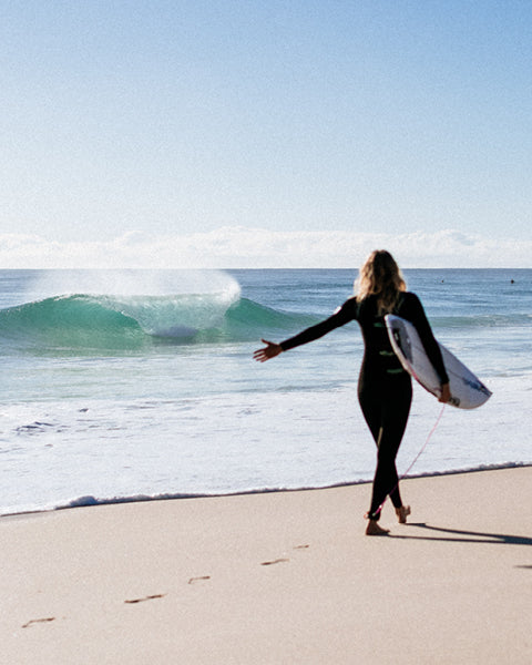 Person in a wetsuit holding a surfboard on a beach with waves in the background