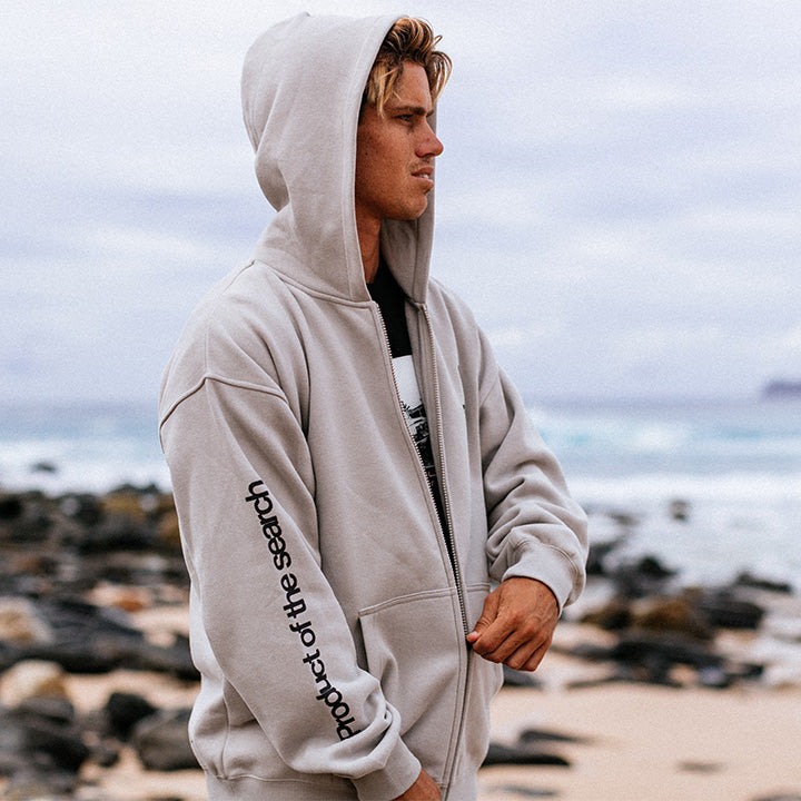 Person wearing a light gray hoodie with text on the sleeve, standing on a rocky beach.