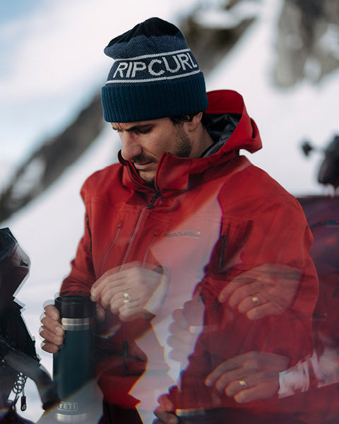 Man wearing a red jacket and blue beanie with 'Rip Curl' logo, holding a black mug outdoors.
