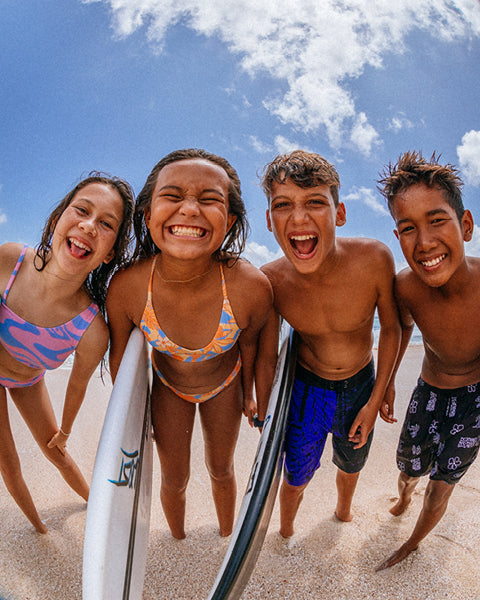 Four children on a beach with surfboards, smiling and laughing under a blue sky.