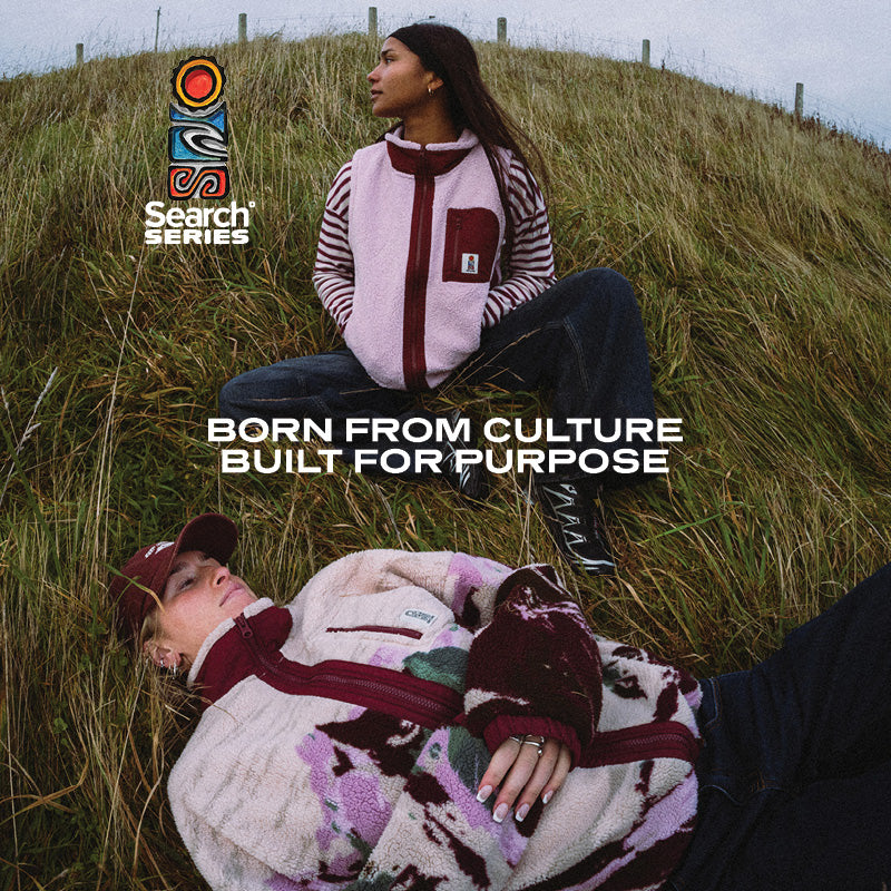 Two models wearing Search Series outerwear are posed on a grassy hillside. One model sits up wearing a light pink and dark red striped fleece vest over a striped long-sleeve shirt and dark jeans, while the other model lies down wearing a fuzzy cream, pink, and dark red patterned fleece jacket and a matching cap.
