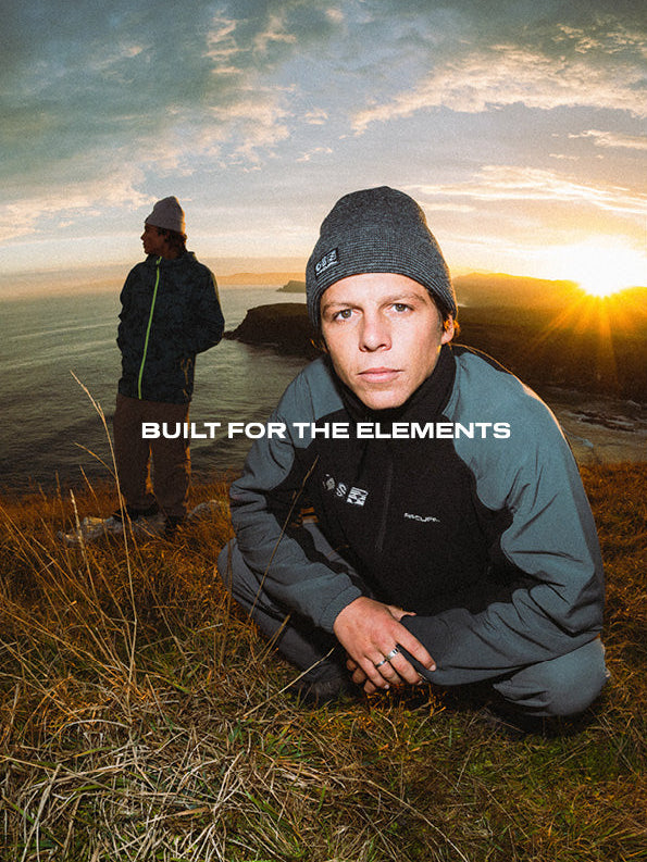 A man in outdoor apparel kneels in grassy terrain at sunset while another person stands in the background by the water. The text 'BUILT FOR THE ELEMENTS' is overlaid across the center.