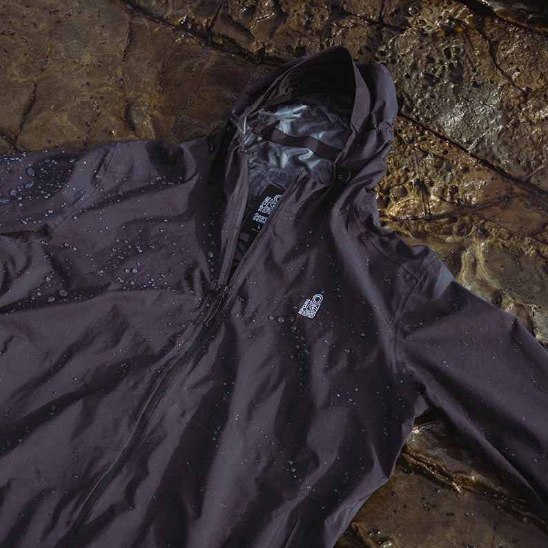 A dark waterproof hooded jacket with water droplets on the surface, laid flat on a wet rocky surface. The jacket has a small white logo on the left chest.
