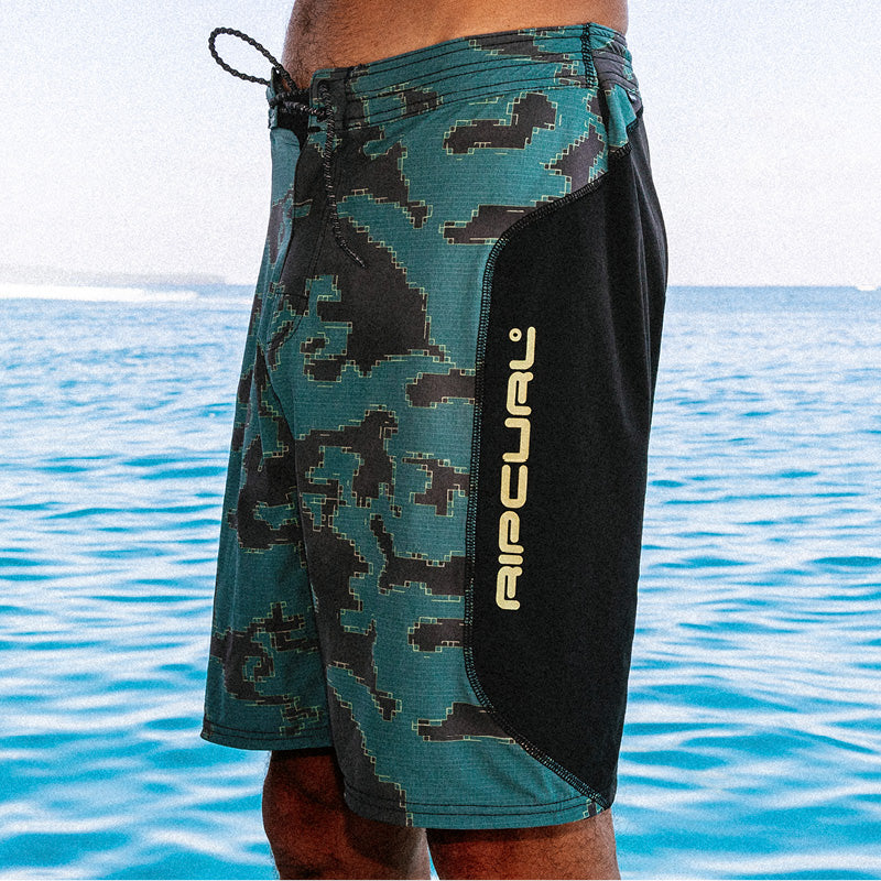 A close-up of a person wearing Rip Curl board shorts with a teal, black, and gray pixelated camouflage pattern and a solid black side panel with the Rip Curl logo, set against a blue ocean background.