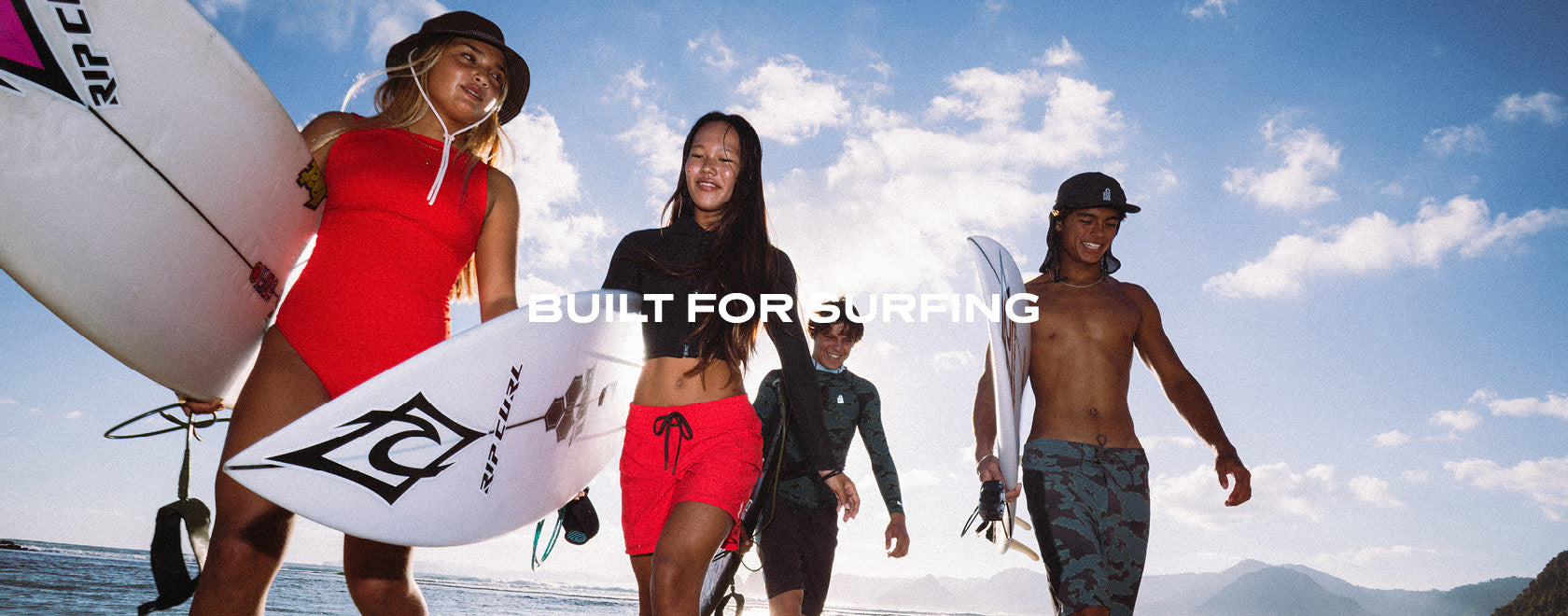 Four surfers walking outdoors on a beach under a blue sky with white clouds. Two women on the left carry white surfboards, one wearing a red one-piece swimsuit, and another in a black top and red shorts.