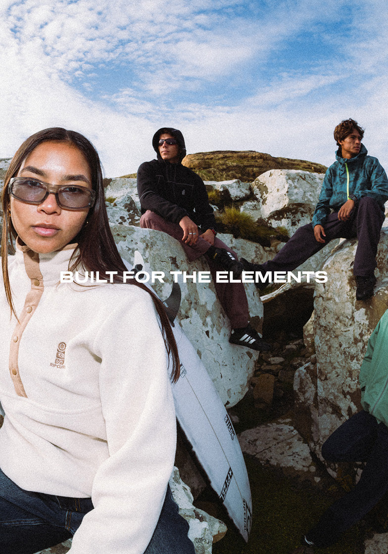 Outdoor lifestyle photo featuring three surfers sitting on white rocks under a blue sky, with the text 'BUILT FOR THE ELEMENTS' overlaid across the center.