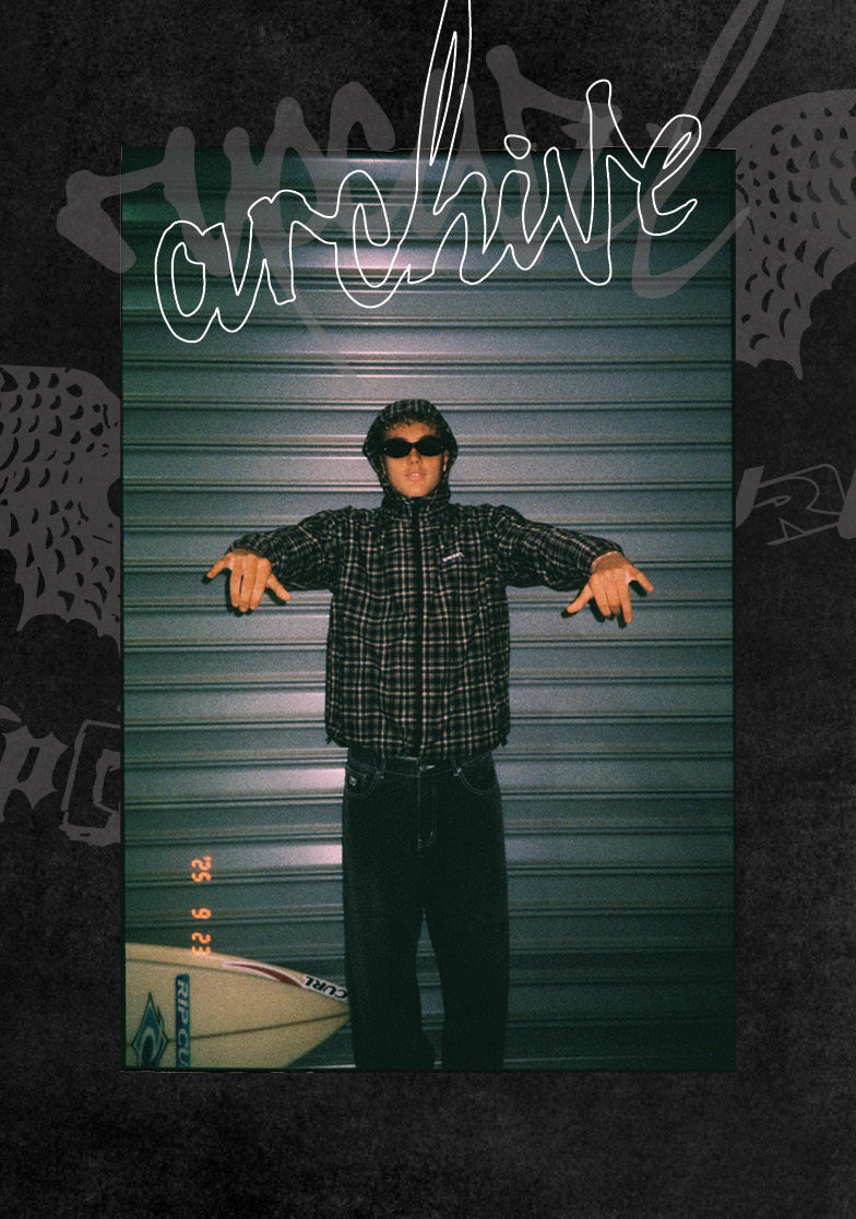 A person wearing a black plaid flannel shirt, black pants, and sunglasses stands with arms outstretched in front of a green metal roll-up door. A white surfboard is partially visible at the bottom left.