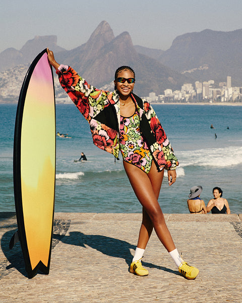 Woman in a colorful outfit holding a surfboard on a beach with mountains and cityscape in the background