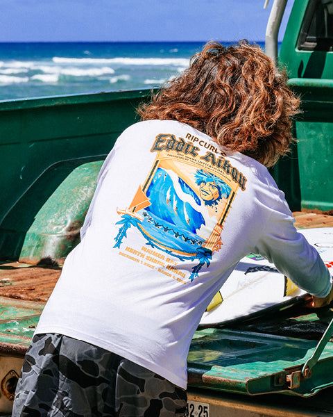 Person wearing a white t-shirt with a graphic design, standing near a green vehicle by the ocean.